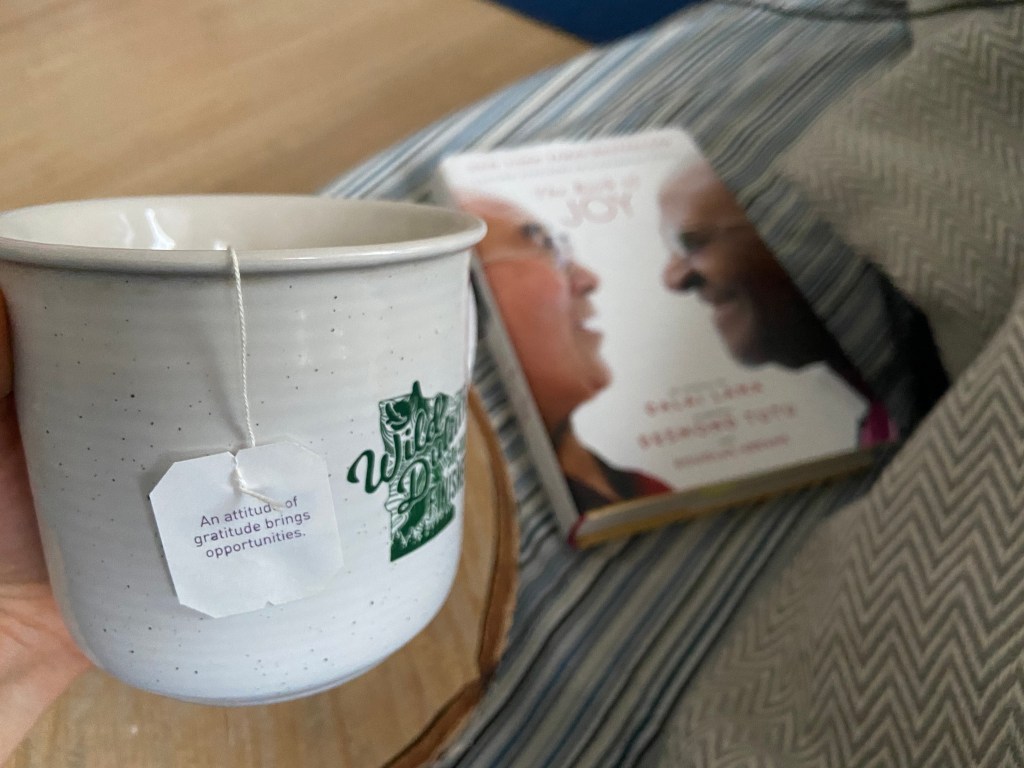 A hand holding a mug of tea, with The Book of Joy in the background