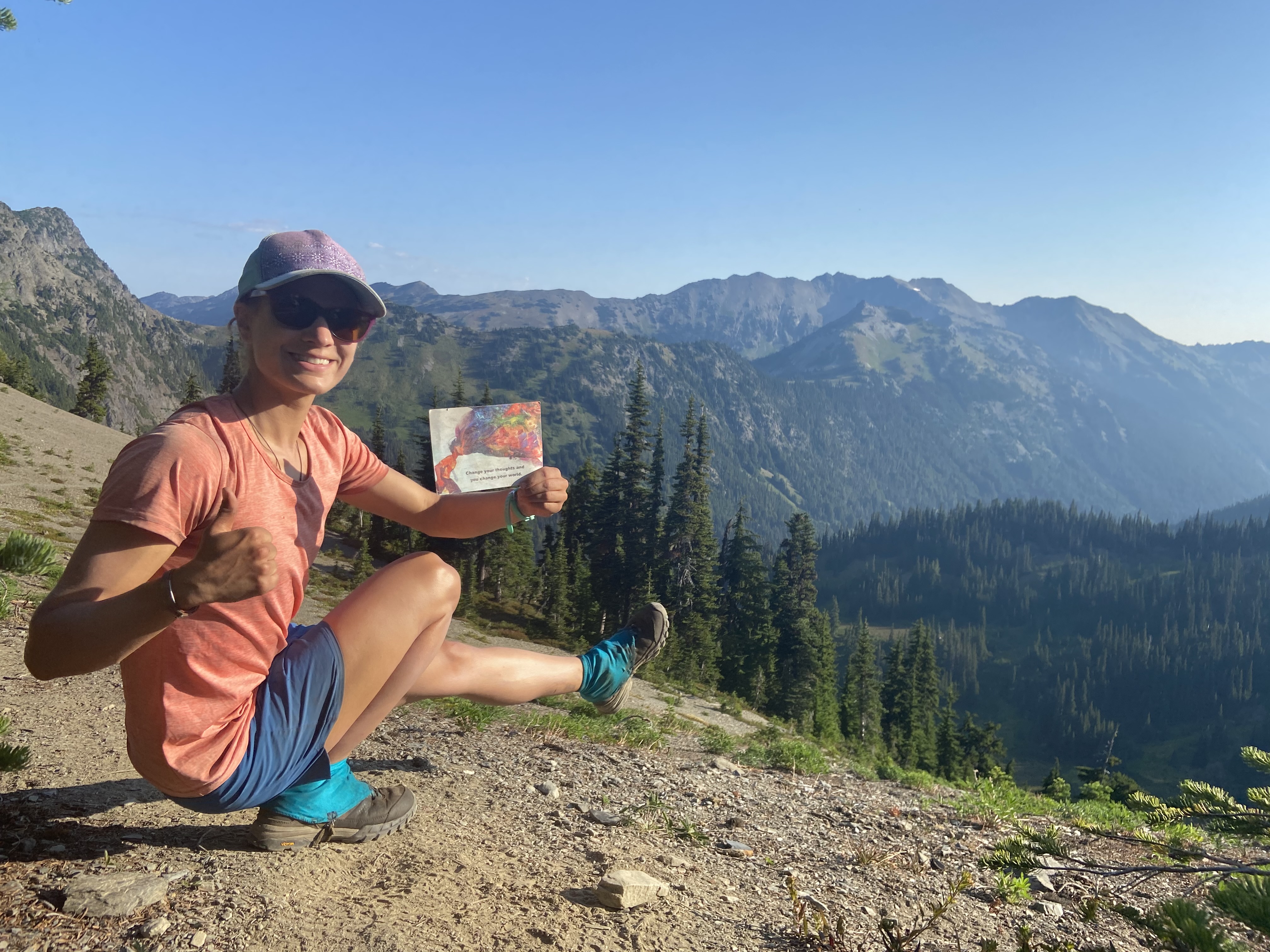 A woman doing a one-legged squat, with mountains in the background and holding a postcard that says "Change your thoughts and you change your world."