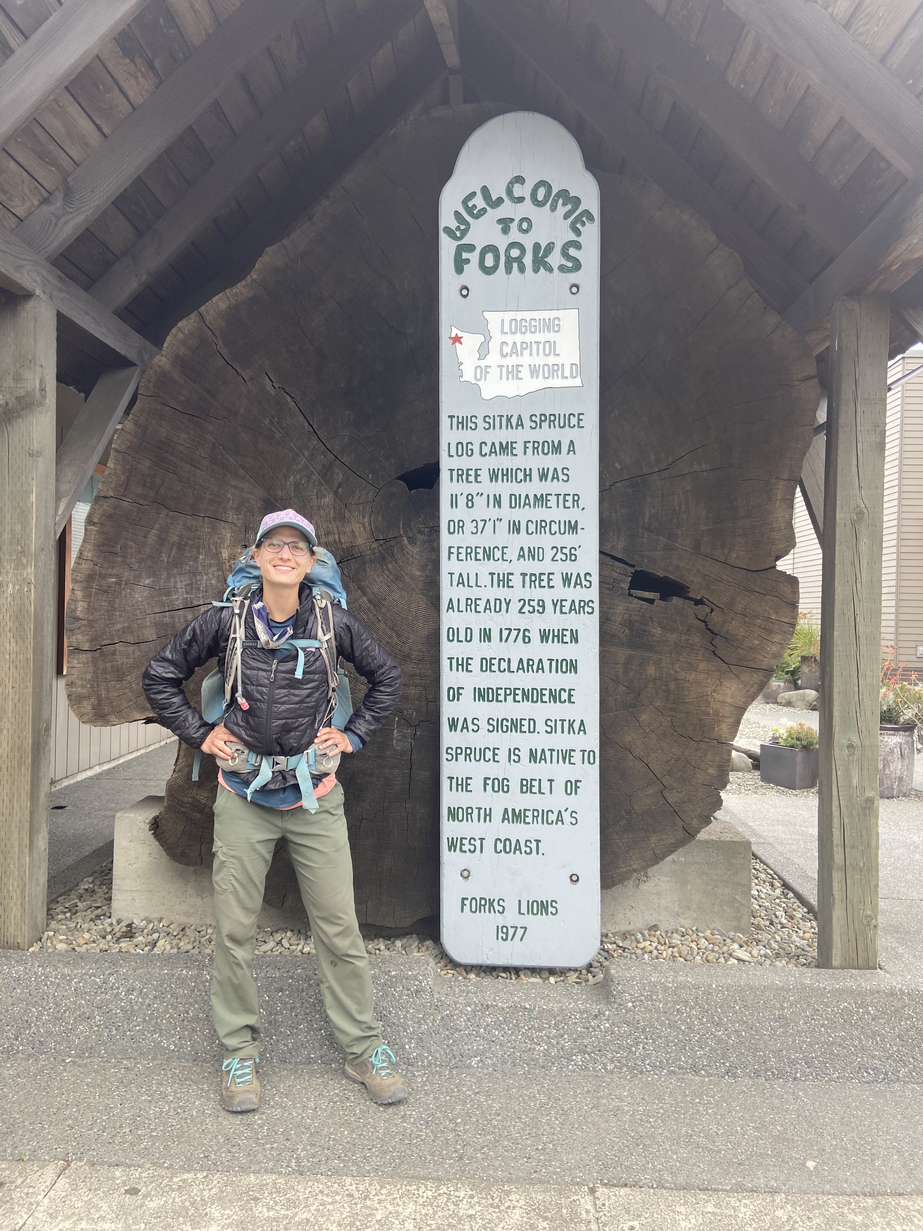 A woman standing in front of a large cut log with a "Welcome to Forks" sign in front of it. The sign says "Welcome to Forks, logging capital of the world. This sitka spruce log came from a tree which was 8 feet in diameter or 37 feet in circumference and 256 feet tall. The tree was already 250 years old in 1776 when the Declaration of Independence was signed. Sitka spruce is native to the fog belt of North America's West Coast. Forks Lions 1977."