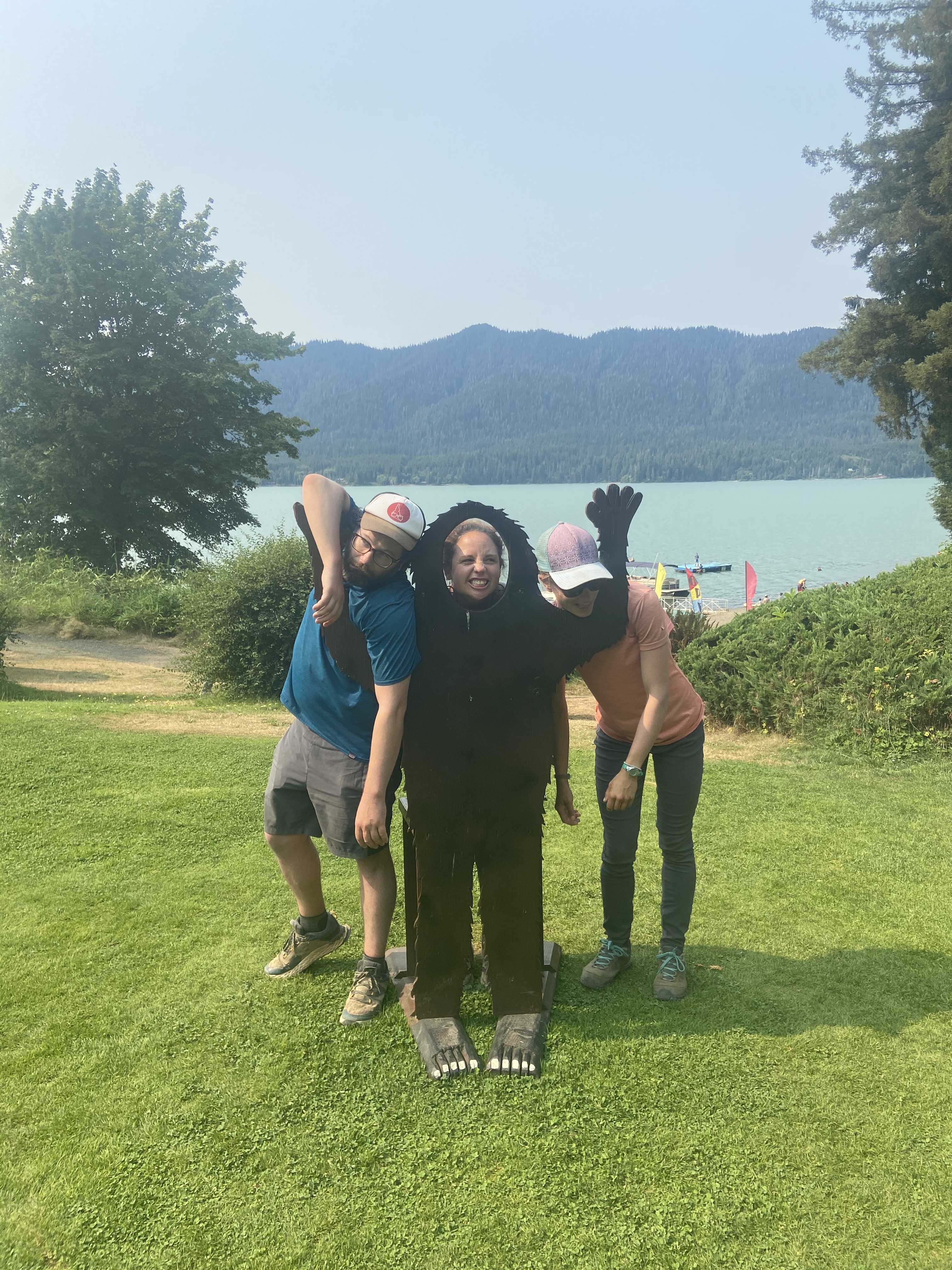 Three people with a lake in the background. One is behind a cutout of bigfoot with their face showing through a cut hole, the other two are pretending to be "caught" in the arms of the Big Foot and are pretending to hang limply at the sides.