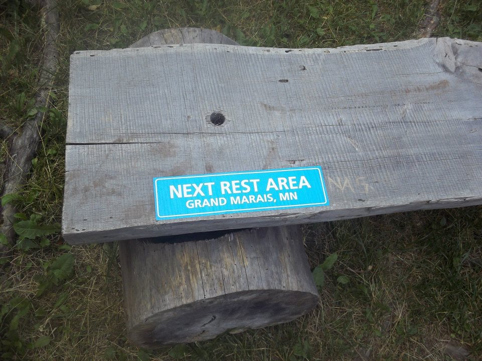 A picture of a wooden bench with a sign that says "Next Rest Area Grand Marais, MN"