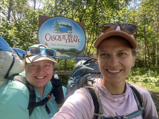 A selfie of two women with backpacks. Behind them a sign says "Casque Isle Trail" Hike the North Shore!"