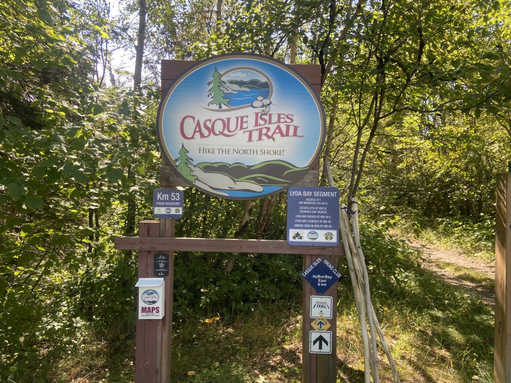 A series of signs. One large sign says "Casque Isle Trail: Hike the North Shore!" Smaller signs say: "Km 53 from Rossport", "Lyda Bay Segment", "Casque Isle Trail Hiking Club", "Voyageur Trail", "Great Trail"; There is also a small container of maps and a few arrows pointing down the trail.