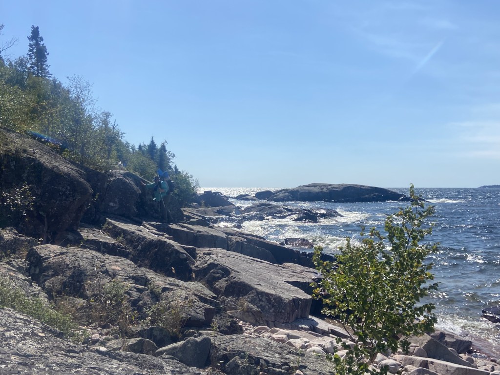 A backpacker traversing a slanted rocky section on the shore.