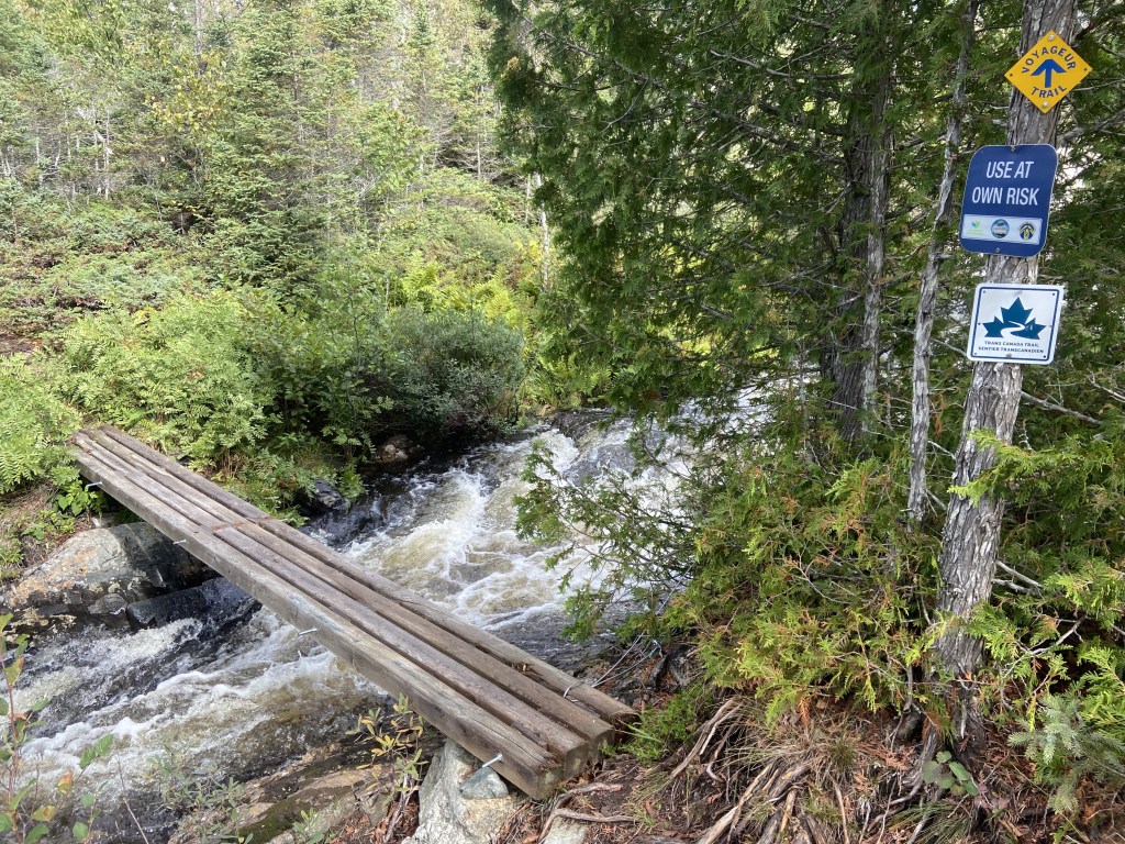 A narrow bridge without railings at a river crossing. A sign that says "use at your own risk".